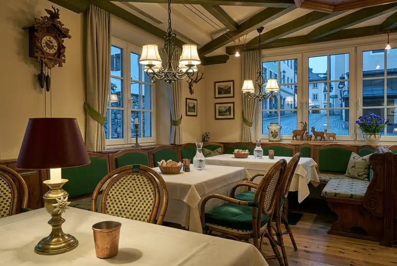 Restaurant Martha Gemütlicher Gastraum eines traditionellen Restaurants mit Holzbalkendecke, Kronleuchtern und mehreren gedeckten Tischen mit weißen Tischdecken. Entlang der Fenster stehen grün gepolsterte Sitzbänke und Holzstühle. An den Wänden hängen Bilder und eine Kuckucksuhr.