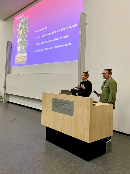 Eine Frau und ein Mann stehen in Universitätsräumen bei einem Vortrag an einem Pult. Im Hintergrund befindet sich eine große Tafel.