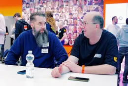 Markus Buff (+Pluswerk Magdeburg buff.rocks) und +Pluswerk CEO Jens Krumm im Gespräch beim TYPO3Camp Mitteldeutschland 2025 – entspannte Stimmung vor einer Fotowand mit buntem Community-Mosaik.