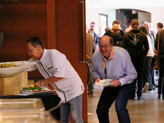 A cheerful attendee at the WebCamp Venlo 2025 buffet, eagerly waiting for a meal while the chef prepares fresh food. In the background, participants are engaged in conversations.