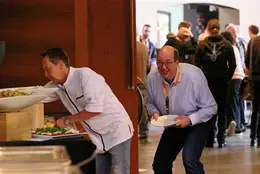 A cheerful attendee at the WebCamp Venlo 2025 buffet, eagerly waiting for a meal while the chef prepares fresh food. In the background, participants are engaged in conversations.