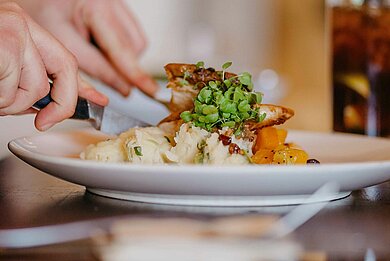 PCCD Dinner – Menü & kulinarische Auswahl Nahaufnahme eines angerichteten warmen Gerichts auf einem Teller in einem Restaurant. Eine Person schneidet das Essen mit Messer und Gabel. Im Hintergrund steht ein Getränk auf dem Tisch.
