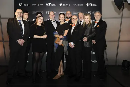 Gruppenfoto während der TYPO3 Award-Gala 2024: Das +Pluswerk-Team und Partner in festlicher Abendgarderobe stehen vor einer Sponsorenwand, einige mit Gläsern in der Hand, lächelnd und feiernd den Erfolg des Abends.