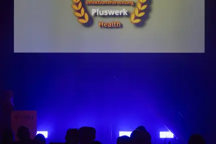 Auf der Leinwand einer Bühne wird der TYPO3 Award 2024 in der Kategorie 'Health' präsentiert, verliehen an das Helmholtz-Zentrum für Infektionsforschung und +Pluswerk. Im Vordergrund sieht man die Silhouetten des Publikums in festlicher Atmosphäre.