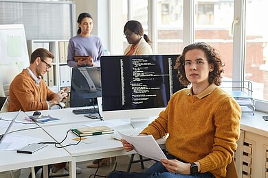 A young software developer wearing glasses and a mustard-colored sweater sits at a desk in a modern office, holding documents. Behind him, a computer screen displays lines of code. In the background, three colleagues collaborate, working on laptops and tablets in a bright, open workspace.