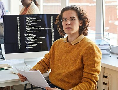 A young software developer wearing glasses and a mustard-colored sweater sits at a desk in a modern office, holding documents. Behind him, a computer screen displays lines of code. In the background, three colleagues collaborate, working on laptops and tablets in a bright, open workspace.