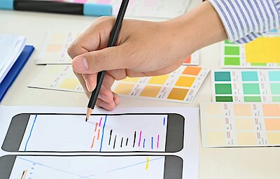 Close-up of a person's hand drawing on a smartphone wireframe layout with a pencil, surrounded by color swatches and design elements. The person is also holding a smartphone, suggesting a focus on mobile app design or user interface development.