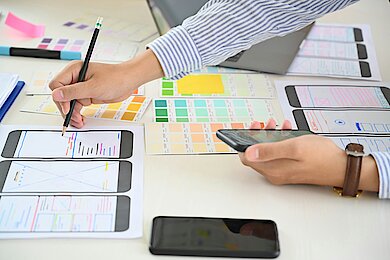 Close-up of a person's hand drawing on a smartphone wireframe layout with a pencil, surrounded by color swatches and design elements. The person is also holding a smartphone, suggesting a focus on mobile app design or user interface development.