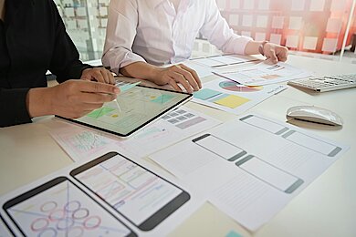 UX/UI-Design-Workshop mit Wireframes und Prototyping Zwei Personen arbeiten an UX- und UI-Designs mit Tablet und ausgedruckten Wireframes.