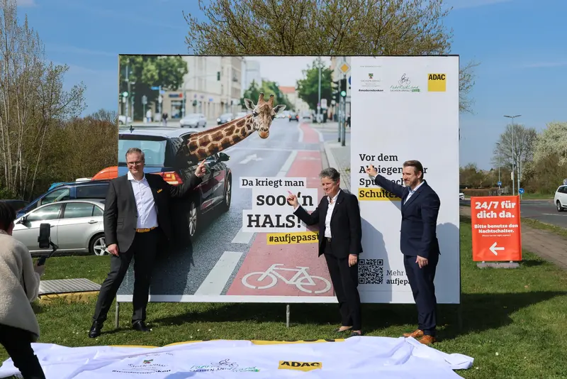 Pressetermin zum Start der Kampagne #aufjepasst in Magdeburg Drei Vertreter der Kampagne #aufjepasst präsentieren bei einem Pressetermin in Magdeburg ein großformatiges Kampagnenmotiv mit einer Giraffe am Autofenster und dem Slogan „Da kriegt man sooo’n HALS!“.