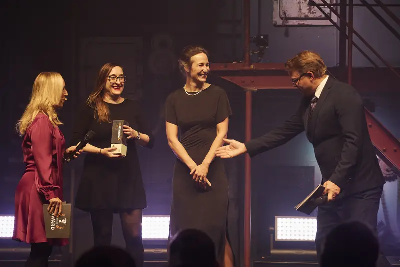 Anna Stephan vom +Pluswerk Standort Dortmund (Mitte links) und Sonja Ziegenbalg vom +Pluswerk Standort Dresden (Mitte rechts) nehmen für das +Pluswerk den TYPO3 Award im Rahmen der TYPO3 Conference Gala entgegen. Links überreicht Moderatorin Emily Whigham mit Mikrofon den Preis, während Moderator Matt Beadle rechts applaudierend zur Gratulation ansetzt.