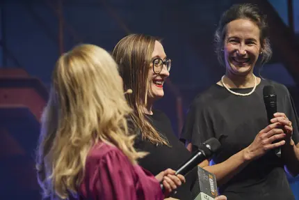 ine Vertreterin von +Pluswerk hält den TYPO3 Award 2024, während sie auf der Bühne mit einer Moderatorin und einer weiteren Person spricht. Die Szene ist von festlicher Atmosphäre und Fokus auf die Preisträgerin geprägt.