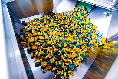 PET-Produktion: Verschlüsse in der Fertigung Große Menge gelb-grüner Kunststoffverschlüsse in einer industriellen Produktionsanlage für PET-Verpackungen, unsortiert in einem Metallbehälter.