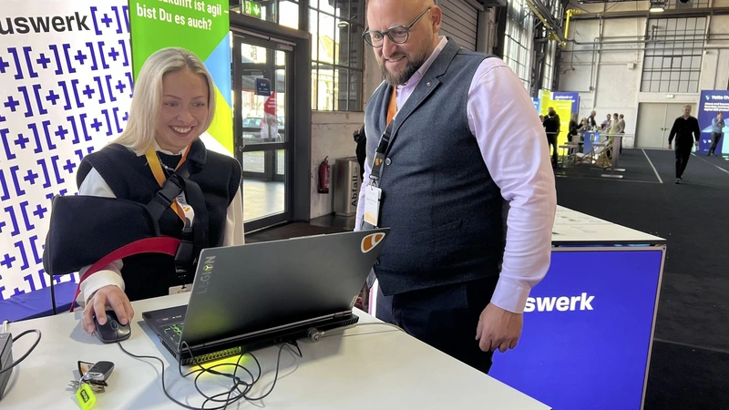 Zwei Personen stehen an einem Tisch am +Pluswerk-Stand auf der TYPO3 Conference 2024. Eine Frau bedient lächelnd eine Computermaus, während ein Mann daneben steht und auf den Laptop blickt. Im Hintergrund sind der +Pluswerk-Banner und Teile des Konferenzgeländes zu sehen. Die Szene vermittelt eine produktive und freundliche Atmosphäre.