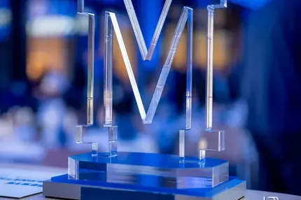 Ein transparentes "M"-Logo aus Acrylglas, das stilvoll von blauer Beleuchtung umrahmt wird. Es steht auf einem eleganten Sockel und symbolisiert den Marketing Award Sachsen-Anhalt 2024.