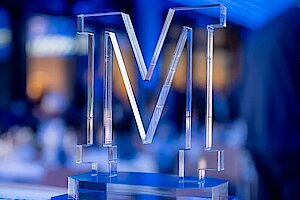 Ein transparentes "M"-Logo aus Acrylglas, das stilvoll von blauer Beleuchtung umrahmt wird. Es steht auf einem eleganten Sockel und symbolisiert den Marketing Award Sachsen-Anhalt 2024.