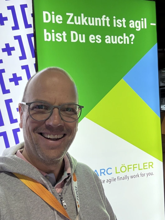 Ein lächelnder Mann mit Konferenzbadge vor einem bunten Hintergrund am +Pluswerk-Stand auf der TYPO3 Conference 2024. Im Hintergrund sind die Worte „Die Zukunft ist agil – bist Du es auch?“ auf einem grünen und blauen Banner zu sehen, flankiert von dem +Pluswerk-Logo.