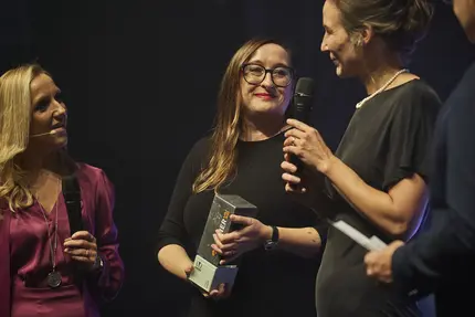 Zwei Vertreterinnen von +Pluswerk lächeln auf der Bühne der TYPO3 Award-Gala 2024, während sie den Gewinner-Pokal entgegennehmen. Eine Moderatorin in violettem Kleid hält ein Mikrofon, während die Szene vor einer industriellen Kulisse stattfindet.