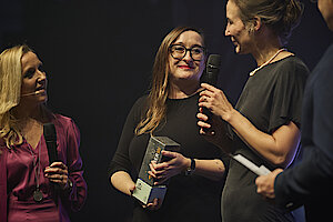 Zwei Vertreterinnen von +Pluswerk lächeln auf der Bühne der TYPO3 Award-Gala 2024, während sie den Gewinner-Pokal entgegennehmen. Eine Moderatorin in violettem Kleid hält ein Mikrofon, während die Szene vor einer industriellen Kulisse stattfindet.