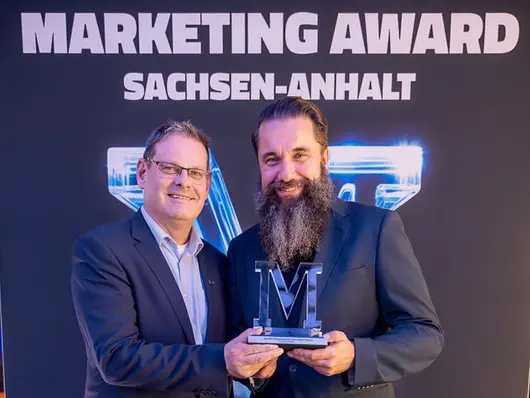 Zwei Männer in Anzügen präsentieren stolz den Marketing Award Sachsen-Anhalt 2024. Sie stehen vor einer Wand mit dem Schriftzug „Marketing Award Sachsen-Anhalt“. Die Trophäe, ein „M“-förmiges Acryl-Logo, wird in die Kamera gehalten.