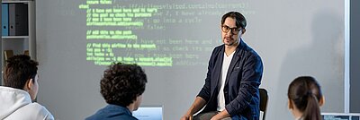 KI-Workshop zur Analyse von Website-Struktur und Code Ein Referent erklärt vor einer projizierten Codeansicht die Struktur einer Website, während drei Teilnehmende an einem Tisch mit Laptops zuhören.