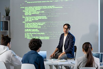 KI-Workshop zur Analyse von Website-Struktur und Code Ein Referent erklärt vor einer projizierten Codeansicht die Struktur einer Website, während drei Teilnehmende an einem Tisch mit Laptops zuhören.