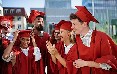 Pluswerk-Hochschulstudie-2024-Studierende-Bild Happy graduates in red gowns stand in a group and talk