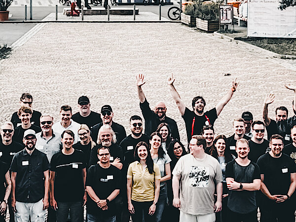 Die +Pluswerk-Crew am Main Die Mitarbeiter von +Pluswerk stehen im Freien am Main-Ufer in Rüsselsheim, winken fröhlich in die Kamera und grüßen zu einem Gruppenfoto.