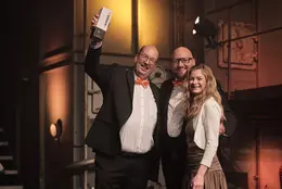 Drei Mitglieder des Earlybird- und +Pluswerk-Teams posieren auf der Bühne mit der TYPO3-Auszeichnung in den Händen und strahlen vor dem Hintergrund der industriellen Beleuchtung.