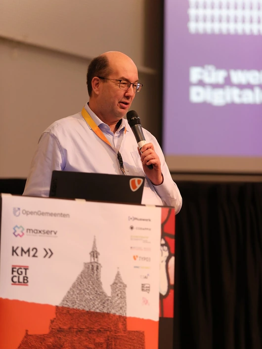 Jens Krumm, CEO of Pluswerk, speaking at WebCamp Venlo 2025. He stands behind a podium with event sponsor logos, delivering his talk with a microphone in hand.