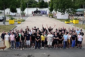 Gruppenfoto von etwa 60 Teilnehmern des Pluscamps 2025, aufgenommen im Freien auf einem gepflasterten Platz in Rüsselsheim mit Blick auf den Main. Die Personen stehen dicht beieinander, einige heben die Arme jubelnd in die Luft. Im Hintergrund ist ein Reiterstandbild, ein „RÜSSELSHEIM“-Schriftzug und viel Grün zu sehen. Ein Hund steht links am Bildrand.