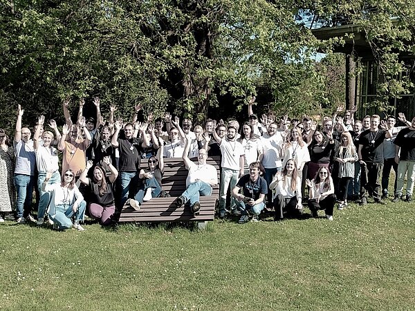 Die +Pluswerk Crew in Karlsruhe beim +Pluscamp Die Mitarbeiter von +Pluswerk stehen im Grünen in Karlsruhe, winken fröhlich in die Kamera und grüßen zu einem Gruppenfoto.