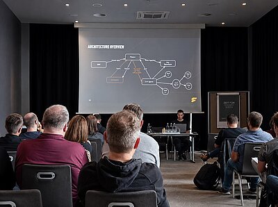 PCDD 2025: Technische Sitzung bei den Pimcore Community Dev Days Die Teilnehmer verfolgen einen Fachvortrag während der Pimcore Community Dev Days 2025 in Frankfurt, während ein Referent auf einer großen Leinwand ein Softwarearchitekturdiagramm erläutert.